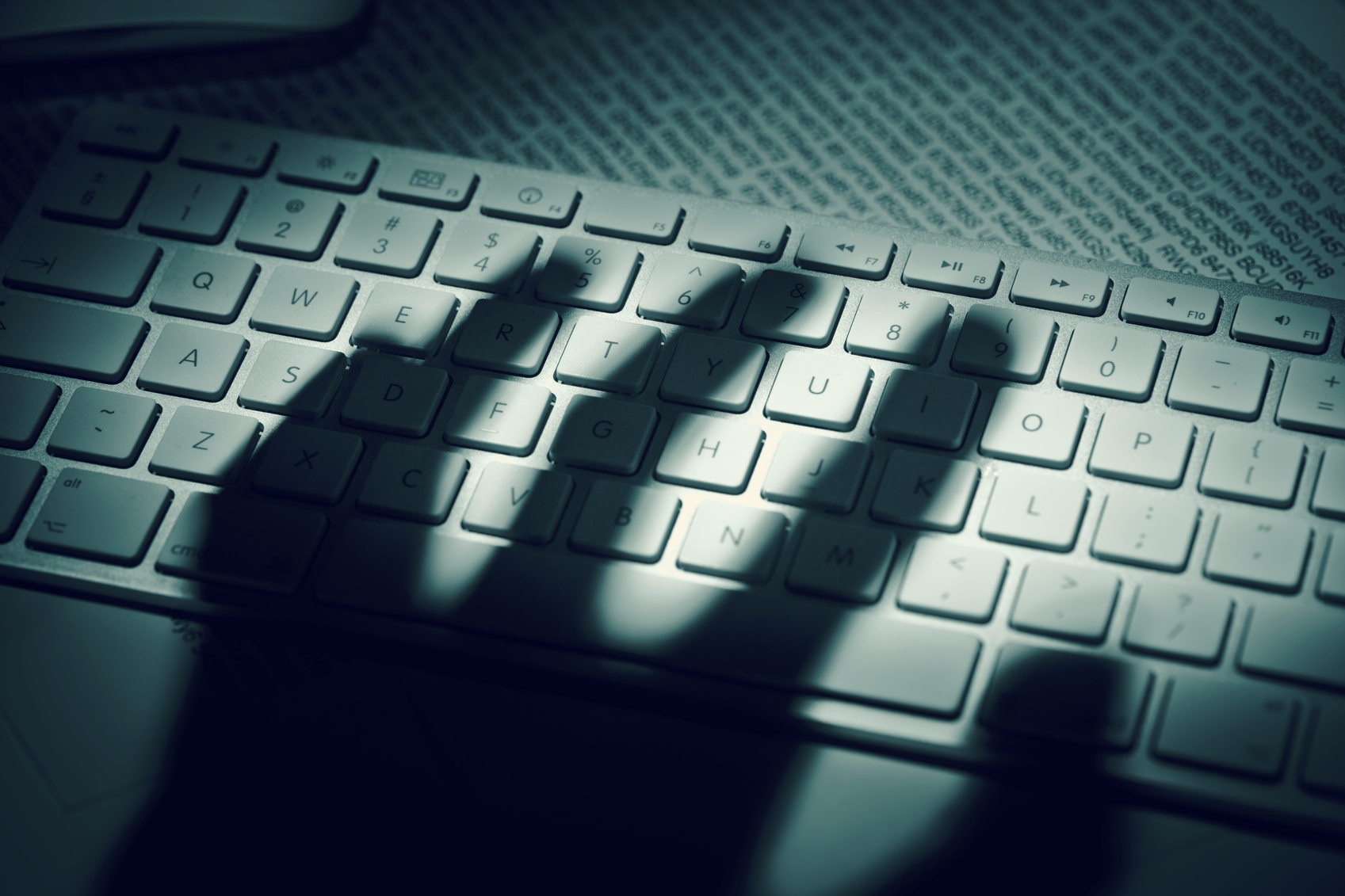 A menacing hand's shadow on a computer keyboard in front of printed computer data. Dramatic light, high contrast.